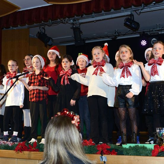 Eine Gruppe junger Mädchen in Weihnachtsoutfits tritt auf einer Bühne mit Mikrofon und Dekorationen auf.