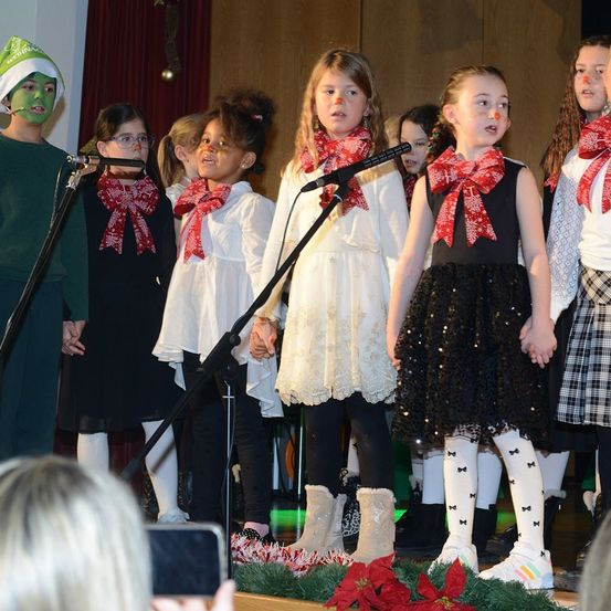 Eine Gruppe von Kindern in Winterkleidung und Stiefeln, die auf der Bühne vor einem Mikrofon singen, mit einem Weihnachtsbaum im Hintergrund.