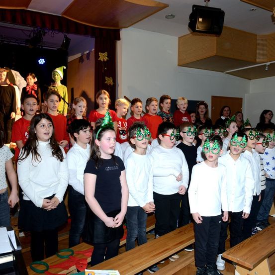 Eine Gruppe von Kindern, gekleidet in Rot und Weiß, mit Weihnachtskopfbändern, steht auf der Bühne und singt. Einige Erwachsene sind im Hintergrund zu sehen, und eine festliche Atmosphäre ist sichtbar.