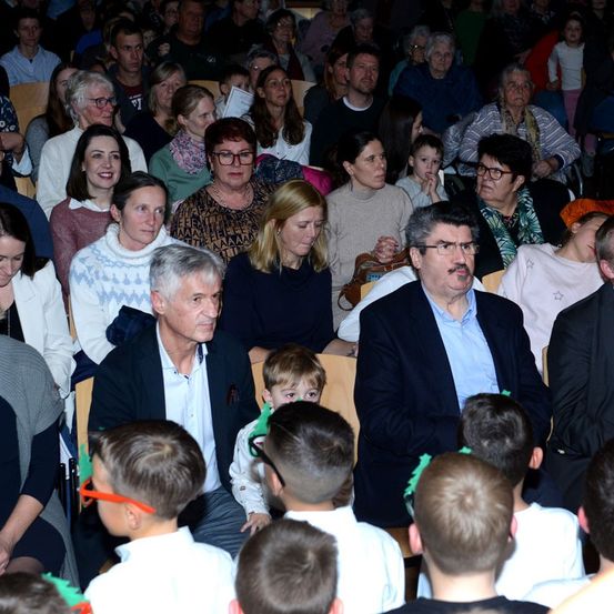 Eine vielfältige Gruppe von Erwachsenen und Kindern sitzt in einem schwach beleuchteten Raum, einige mit Brillen. Zwei Männer sitzen in der ersten Reihe, einer mit einem Schnurrbart und der andere mit einem Kind.