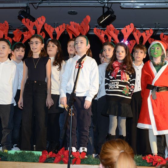 Eine Gruppe von Kindern in Weihnachtskostümen, einschließlich Rentierkopfbändern, tritt auf einer Bühne mit Weihnachtsthema auf.