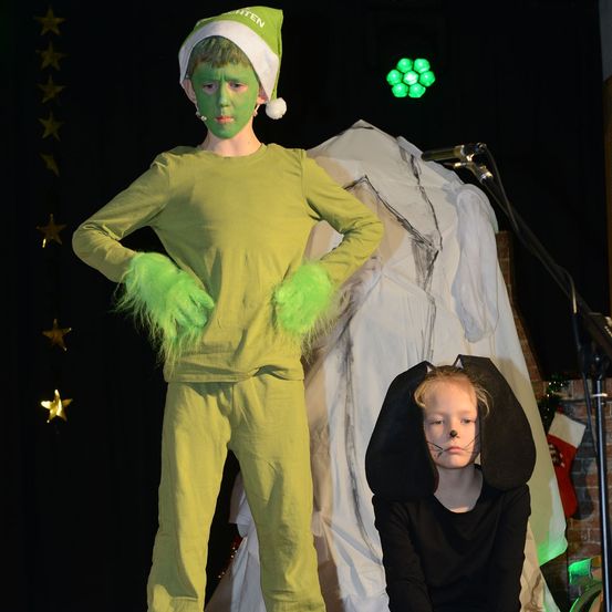 Ein Kind in einer grünen Grinch-Kostümierung steht auf der Bühne mit einem Mikrofon. Daneben steht ein Kind in einem schwarzen Hundekostüm, beide vor einem Hintergrund von Sternen und einem grünen Licht.