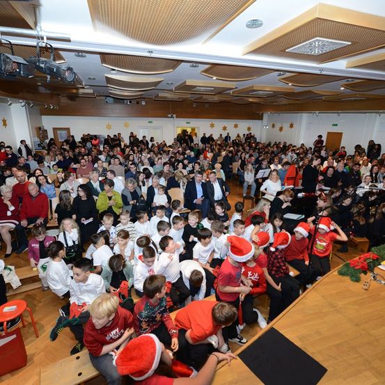 Eine große Gruppe von Menschen in einem Saal, viele mit Weihnachtsmützen, versammelt sich um eine Bühne. Kinder sitzen auf Bänken, während Erwachsene dahinter stehen. Eine Bühne mit Klavier und Weihnachtsdekoration.