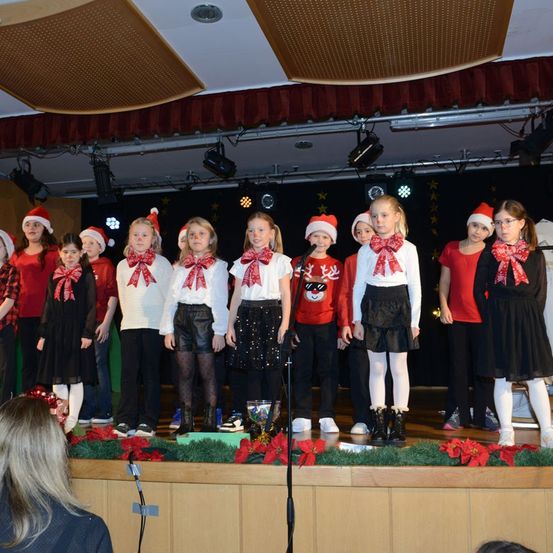 Eine Gruppe von Kindern in Weihnachtskostümen steht auf der Bühne und singt. Die Bühne ist mit roten Blumen und Grünpflanzen geschmückt. Die Kinder tragen passende rote und weiße Outfits mit Weihnachtsmützen. Eine Frau beobachtet sie von unten.