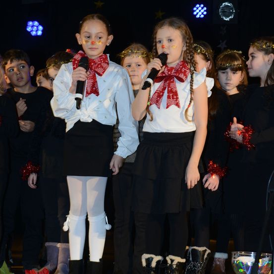 Zwei junge Mädchen in festlichen Kostümen und Stiefeln singen in Mikrofone, stehend unter anderen Kindern, möglicherweise bei einer Feier.