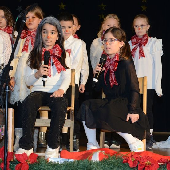 Eine Gruppe von Kindern auf der Bühne, mit roten Schleifen und Mikrofonen, eine Mädchen in einem schwarzen Kleid singt.
