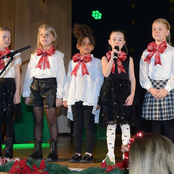 Fünf junge Mädchen stehen auf einer Bühne mit Mikrofonen vor einem Publikum. Sie tragen passende rote Schleifen und singen wahrscheinlich ein Weihnachtslied.