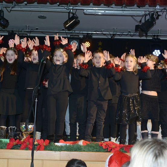 Eine Gruppe von Kindern in schwarzer Kleidung tritt auf einer mit roten Weihnachtssternen und einem Mikrofon geschmückten Bühne auf. Sie haben festliche Stirnbänder und scheinen zu singen.