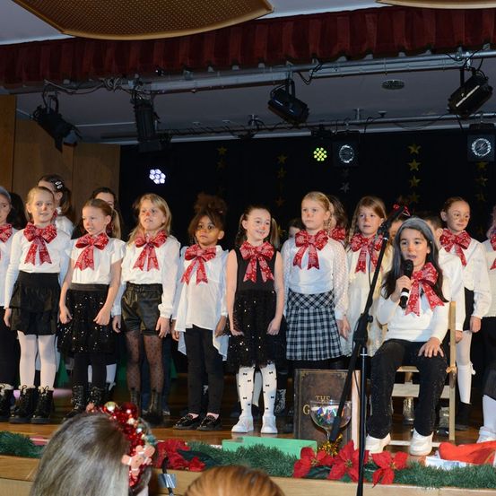 Eine Gruppe junger Mädchen, gekleidet in identischen weißen Oberteilen und Röcken, tritt auf einer Bühne auf, geschmückt mit roten Schleifen und Mikrofonen. Sie sind von Bühnenbeleuchtung und Dekorationen umgeben.