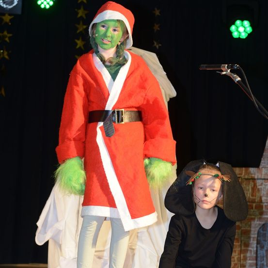 Ein junges Mädchen, verkleidet als Grinch, steht auf der Bühne, während ein weiteres Kind, verkleidet als Hund, vor ihr kniet. Hinter ihnen ist ein Hintergrund mit gelben Sternen zu sehen.
