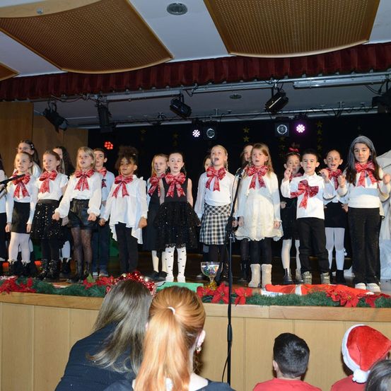 Eine Gruppe von Kindern tritt auf der Bühne auf, mit Schleifen um den Hals, in weißen und schwarzen Outfits. Sie singen vor einem Publikum mit Weihnachtsthema.