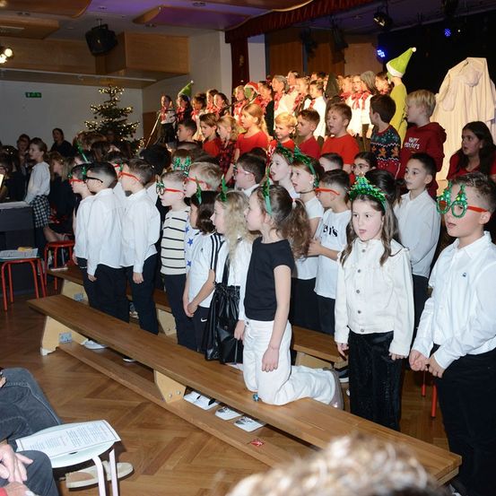 Kinder in festlicher Kleidung singen gemeinsam in einem Raum mit einem Weihnachtsbaum, einer Bühne und einer weißen Figur im Hintergrund.