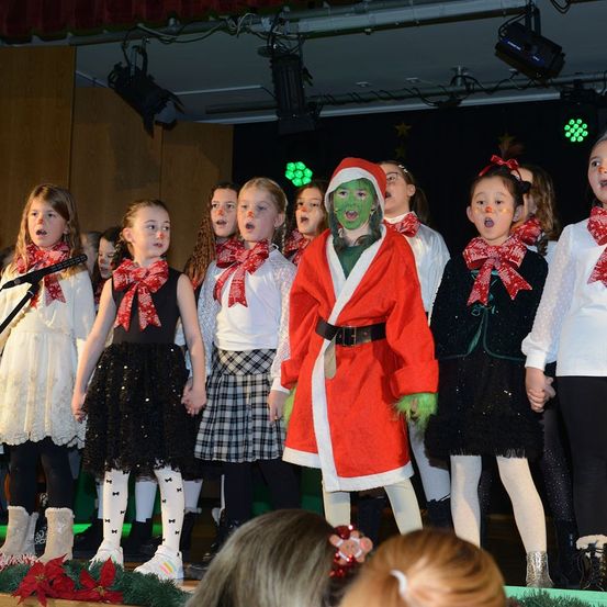 Eine Gruppe von Kindern in festlicher Kleidung, darunter eine als Grinch verkleidet, tritt auf einer Bühne mit Weihnachtsdekorationen auf.