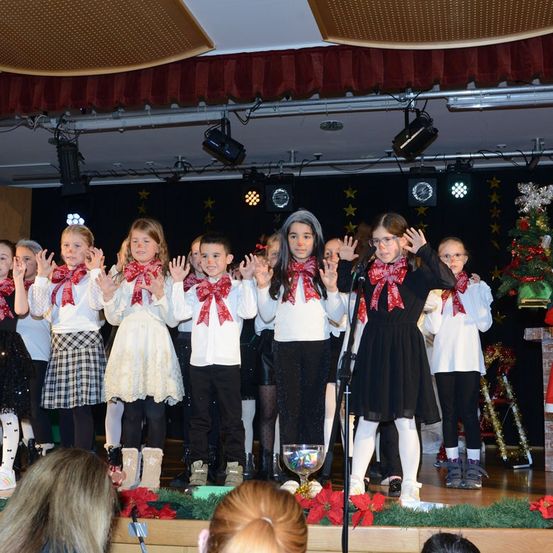 Eine Gruppe von Kindern tritt auf der Bühne auf, gekleidet in passende Outfits mit roten Schleifen. Sie stehen vor einem Mikrofon, und ein Weihnachtsbaum ist im Hintergrund zu sehen.