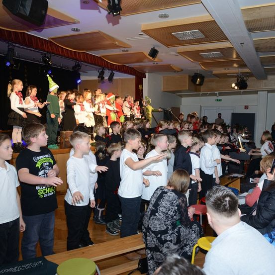 Kinder in verschiedenen Kostümen treten auf einer Bühne auf, während ein Publikum zuschaut. Einige Kinder stehen auf Bänken, andere auf dem Boden, und einige Erwachsene sitzen auf Stühlen.