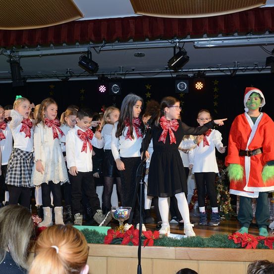 Eine Gruppe von Kindern, die in Weiß mit roten Schleifen und Stiefeln gekleidet sind, singt auf einer Bühne mit einer Frau in Schwarz. Ein Mann, der als Grinch verkleidet ist, steht auch auf der Bühne und zeigt auf die Menge.