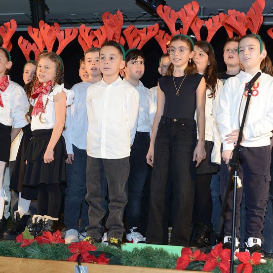 Eine Gruppe von Kindern mit Rentierkopfbändern steht auf einer Bühne vor roten Christsternen. Sie singen, während ein Mikrofon rechts neben ihnen positioniert ist.