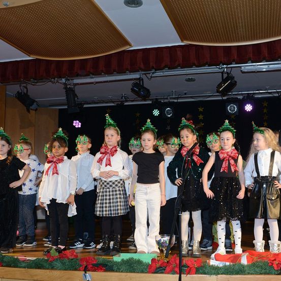 Eine Gruppe junger Kinder in Weihnachtskostümen und Stirnbändern steht auf der Bühne, wahrscheinlich für ein Publikum. Die Bühne ist mit Weihnachtsbäumen und Lichtern dekoriert.