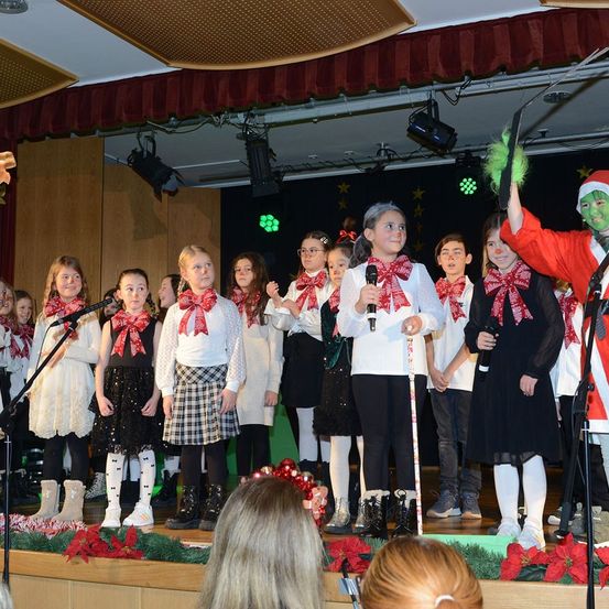 Eine Gruppe junger Mädchen in Weihnachtskostümen tritt auf der Bühne auf. Ein Mensch in einer Grinch-Verkleidung steht rechts und hält ein Mikrofon. Die Bühne ist mit roten Schleifen und Weihnachtsblumen dekoriert.