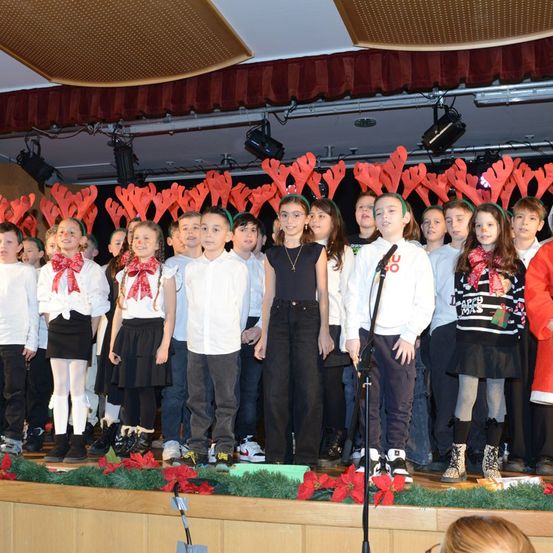 Eine Gruppe von Kindern in Winterkleidung und roten Kopfbändern steht auf der Bühne mit roten Geweihen darüber. Einige tragen Schleifen und Turnschuhe, andere haben Stiefel. Ein Mikrofon ist vorhanden.