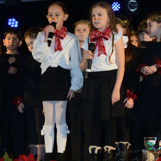 Zwei Mädchen singen auf der Bühne bei einer Schulveranstaltung. Sie halten Mikrofone mit roten Schleifen und tragen weiße Blusen, schwarze Röcke und weiße Strumpfhosen. Hinter ihnen sehen andere Kinder zu, einige mit Kränzen.