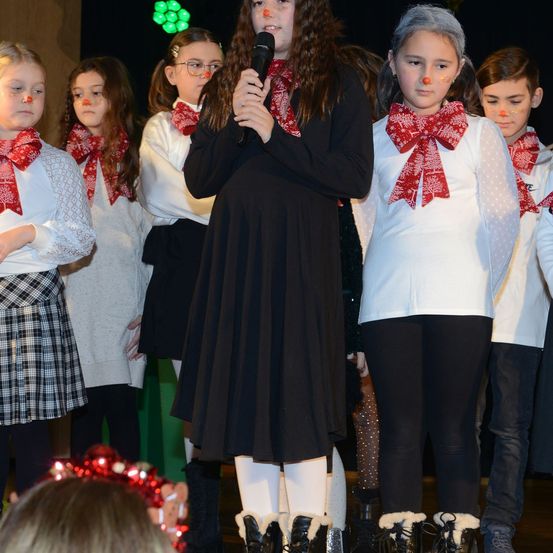 Ein Chor von Kindern, gekleidet in festliche Winterkleidung, tritt auf der Bühne auf. Ein Kind hält ein Mikrofon und singt, während andere hinter ihr stehen. Alle haben rote Schleifen auf ihren Nasen.