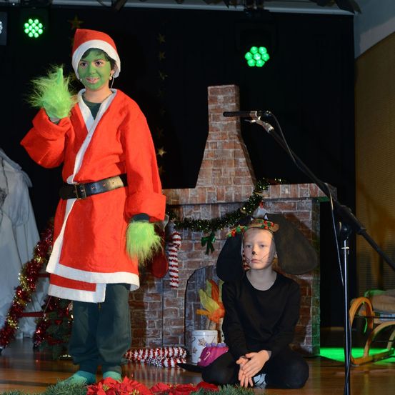 Eine Person in einem Weihnachtsmann-Kostüm mit grüner Gesichtsbemalung steht auf der Bühne, während ein Kind in einem Hundekostüm in der Nähe sitzt. Hinter ihnen steht ein Backsteinkamin mit Weihnachtsdekoration.
