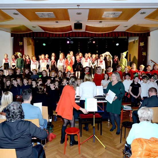 Eine große Gruppe von Kindern und Erwachsenen tritt mit Musikinstrumenten und Dekorationen auf der Bühne auf. Das Publikum sitzt in Stühlen vor ihnen.