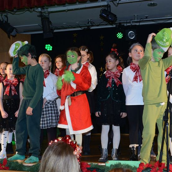 Eine Gruppe von Kindern in Kostümen steht auf der Bühne. Zwei sind als Grinch verkleidet. Ein Junge hält einen grünen Hut, während ein Mädchen ein rotes Santa-Kostüm trägt. Andere tragen rote Schals. Die Bühne ist mit grünen Pflanzen und roten Blumen dekoriert.