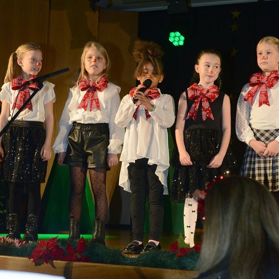 Fünf Mädchen in weihnachtlich geschmückten Outfits stehen auf der Bühne, eines hält ein Mikrofon. Dahinter ist ein grüner Hintergrund mit Sternen und einem Weihnachtsbaum.