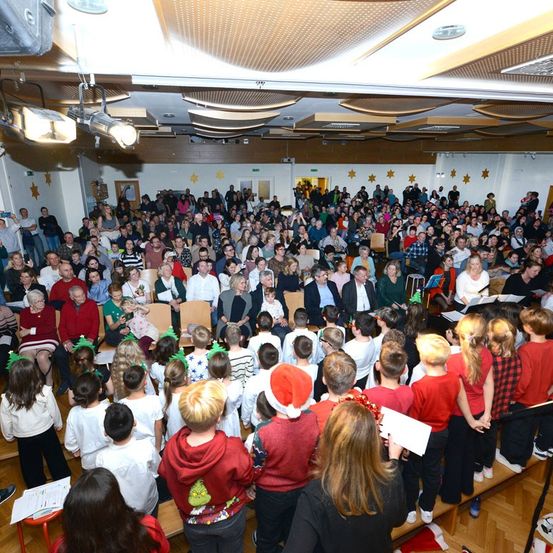 Eine große Menge versammelt sich in einem Saal mit dekorativen Sternen. Viele sind in festlicher Kleidung. Kinder stehen auf der Bühne und singen, während Erwachsene hinter ihnen sitzen oder stehen. Der Saal hat Holzböden und hohe Decken mit Beleuchtungskörpern.
