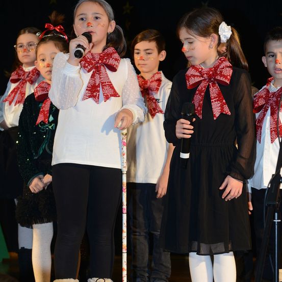Eine Gruppe von Kindern in festlicher Kleidung, mit roten Schals mit weißen Mustern, steht auf einer Bühne. Zwei Mädchen vorne halten Mikrofone, während andere hinter ihnen stehen. Sie scheinen an einer Weihnachtsaufführung teilzunehmen.