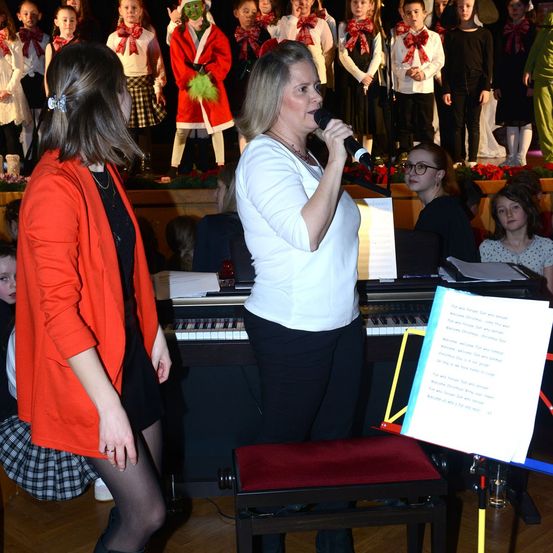 Eine Frau singt in ein Mikrofon, begleitet von einem Pianisten, während ein Chor von Kindern in festlicher Kleidung hinter ihnen steht. Ein Mädchen in einer orangefarbenen Jacke steht neben der Sängerin.