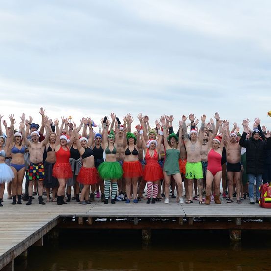 Eine große Gruppe von Menschen, die in Badekleidung und festlichen Mützen gekleidet sind, versammelt sich auf einem Steg am Wasser und hebt ihre Hände in die Luft.