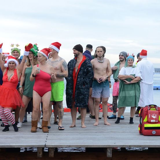 Eine Gruppe von Menschen in festlicher Kleidung, einschließlich Weihnachtsmützen, steht auf einem Steg. Einige sind in Badekleidung, während andere Roben tragen. Eine medizinische Tasche ist in der Nähe.