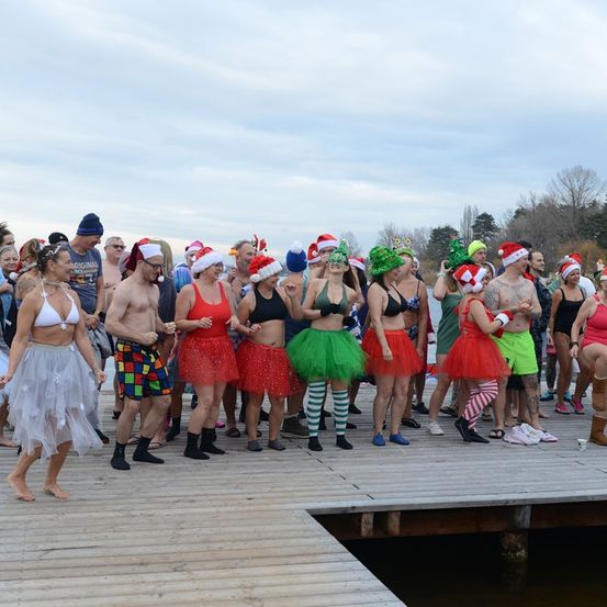 Eine Gruppe von Menschen in festlicher Kleidung versammelt sich auf einem Holzsteg, einige in bunten Tüllröcken und andere in Badekleidung, alle tragen Weihnachtsmützen.