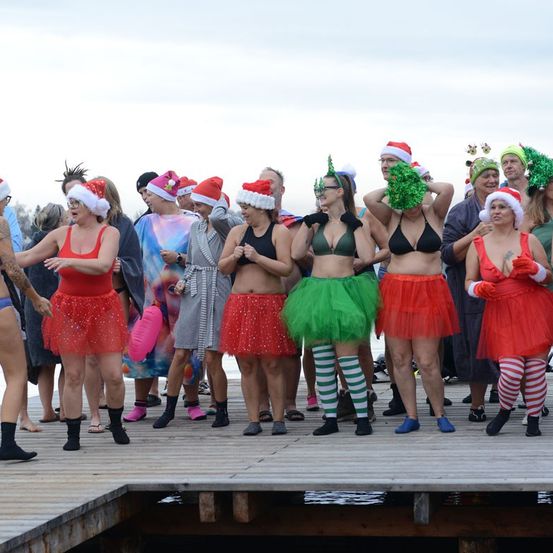 Eine Gruppe von Menschen steht auf einer Holzplattform und trägt festliche Mützen und Kostüme. Einige sind in Bikinis, andere in Kleidern. Sie sind alle barfuß, und das Wasser ist ruhig.