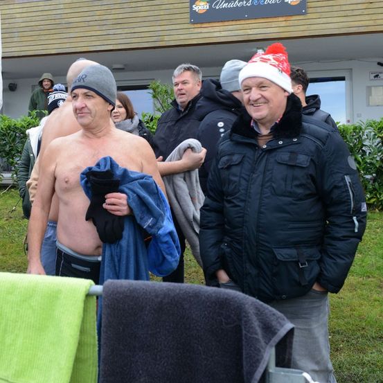 Mehrere Männer stehen draußen in Winterkleidung. Ein Mann ohne Hemd hält ein blaues Handtuch. Ein anderer Mann trägt einen Weihnachtsmannhut. Sie stehen in der Nähe eines Gebäudes mit einem Schild.