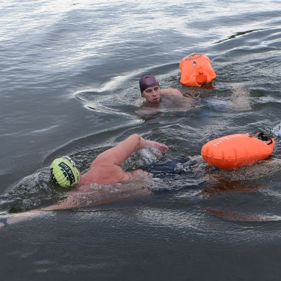 Zwei Schwimmer in einem Gewässer, die Schwimmkappen und Schwimmbrillen tragen. Einer hat einen Rettungsring an sich befestigt.