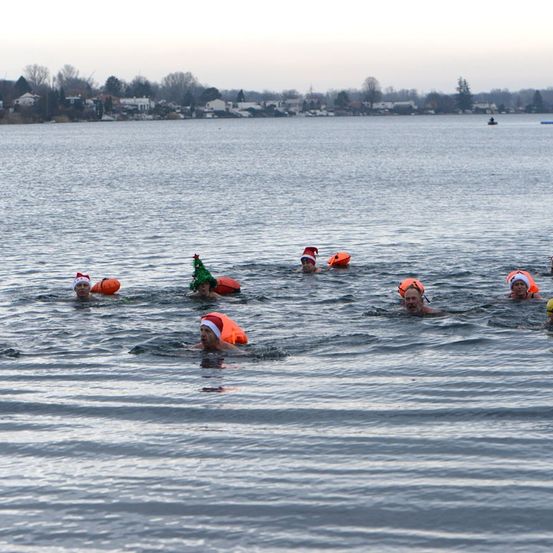 Eine Gruppe von Menschen schwimmt in einem See und trägt Weihnachtsmützen und Schwimmhilfen. Im Hintergrund sind Häuser und ein kleines Boot zu sehen.