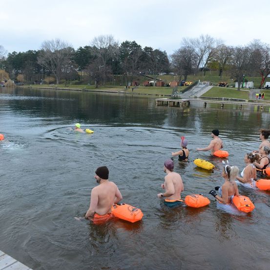 Eine Gruppe von Menschen schwimmt mit Schwimmwesten in einem See. Einige tragen Mützen und Schwimmbrillen. Ein Steg und Bäume sind im Hintergrund zu sehen.