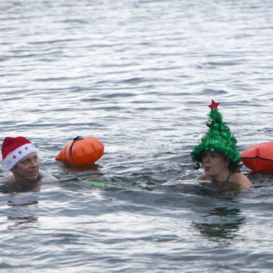 Zwei Personen schwimmen in einem See und tragen Weihnachtsmützen. Einer hat eine Santa-Mütze und der andere eine Weihnachtsbaummütze. Sie sind von schwimmenden Rettungsringen umgeben.