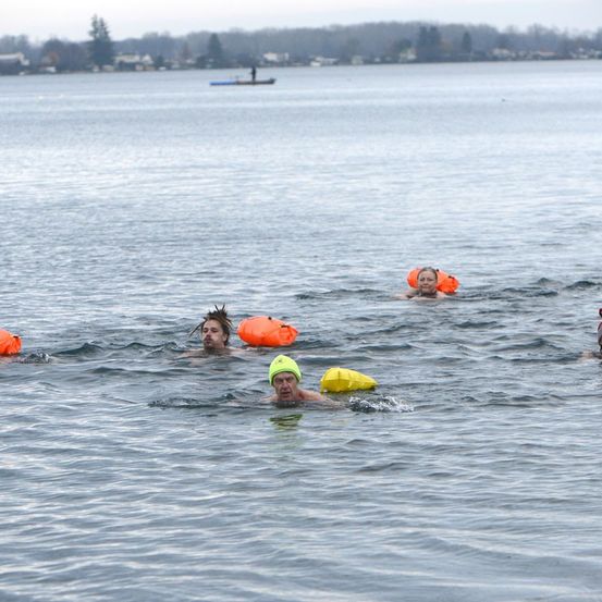 Mehrere Menschen schwimmen in einem See mit Schwimmwesten. In der Ferne ist ein kleines Boot zu sehen.