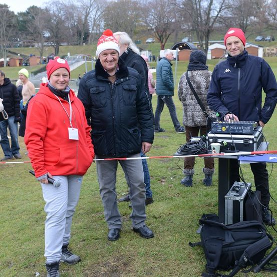 Drei Menschen stehen in einem Grasbereich und tragen Winterkleidung. Sie lächeln und scheinen ein Gespräch zu führen. Hinter ihnen steht ein Mann neben einem Tisch mit Ausrüstung.