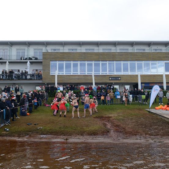 Eine Menschenmenge beobachtet eine festliche Veranstaltung am Fluss. Teilnehmer in Kostümen laufen auf dem Gras, und im Hintergrund befindet sich ein Gebäude mit Glasfenstern.