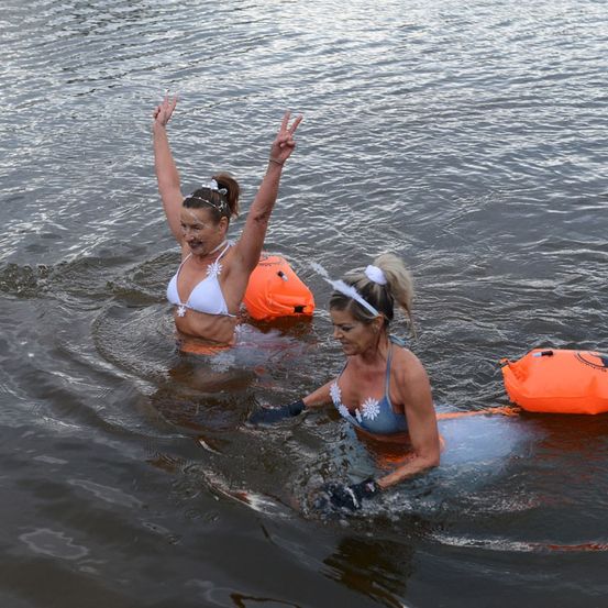 Zwei Frauen schwimmen im Wasser, tragen Badeanzüge und Handschuhe. Eine hat weißes Gesichtsmake-up, die andere ist blau. Sie sind von orangefarbenen Schwimmhilfen umgeben.