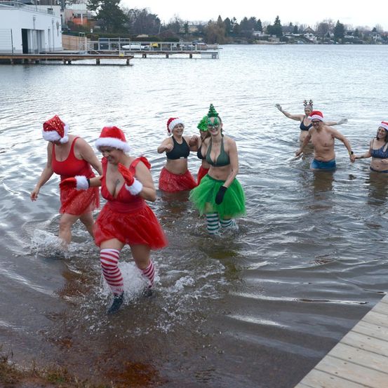 Eine Gruppe von Menschen in Weihnachtsmützen und festlichen Kostümen geht in einen See. Sie nehmen wahrscheinlich an einer Feier teil.