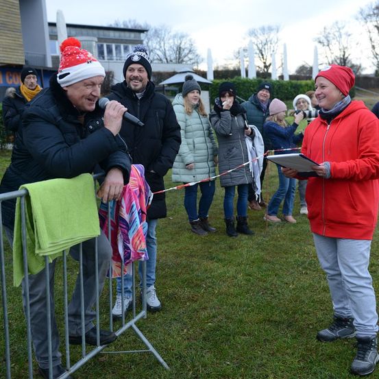 Ein Mann mit Weihnachtsmütze hält ein Mikrofon vor eine Menschenmenge auf einem Rasenfeld. Eine Frau in Rot beobachtet.
