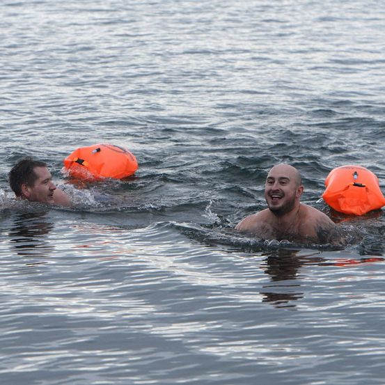 Zwei Männer schwimmen in ruhigem Wasser mit orangefarbenen Schwimmhilfen. Ein Mann lächelt, der andere schaut nach vorne.