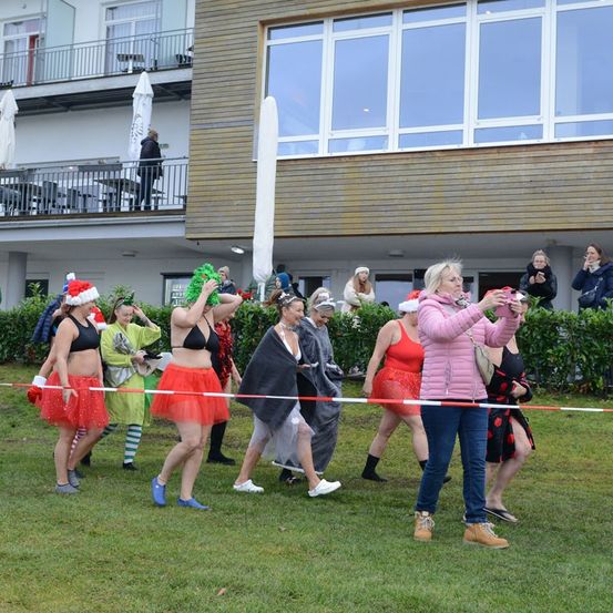 Eine Gruppe von Frauen in festlicher Kleidung, einschließlich Santa-Hüten, tanzt auf einem Feld. Eine Frau in einer rosa Jacke macht ein Foto von ihnen. Hinter den Tänzerinnen befindet sich ein Gebäude mit Glasfenstern und Balkonen.
